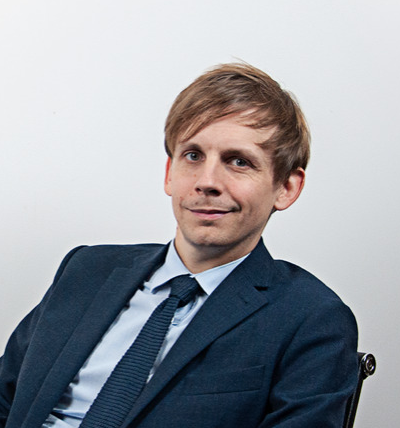 Portrait d'un homme blond au cheveux raides sur fond bland. Il porte un costume bleu gris et une cravate. Il est assis sur une chaise et fait un sourire en coin à la caméra