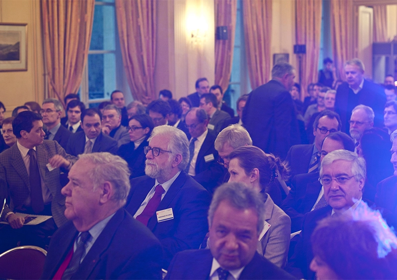 Plusieurs hommes âgés en costumes sont assis dans une salle de conférence. Ils s'installent pour assister à une conférence d'économie. Un halo bleu violet recouvre partiellement l'image.