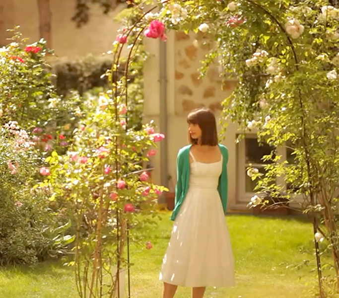 Capture d'une publicité pour n hotel de luxe à Paris. Une jeune femme en robe blanche et gilet vert se promène au milieu d'un jardin très fleuri. Elle est debout et regarde vers la gauche. Vidéo promotionelle tournée par Damien Rougier Réalisateur et motion deisgner